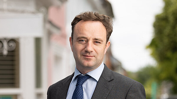 Danny Chambers MP, wearing a dark grey suit and a navy tie. Behind him is a bay window, the exterior is painted white, with pink and blue houses either side