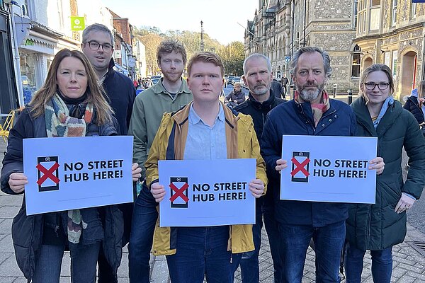 Seven people standing at the east end of Winchester High Street with the Guildhall in the background, three of whom are holding signs saying "No Street Hub here!"